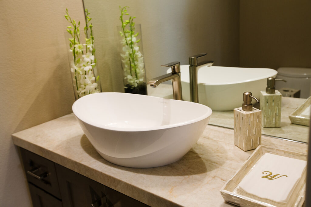 Vessel bowl with a deck mounted brushed nickel single lever faucet on a travertine slab counter top with full height mirror backslash shows contemporary interior design.